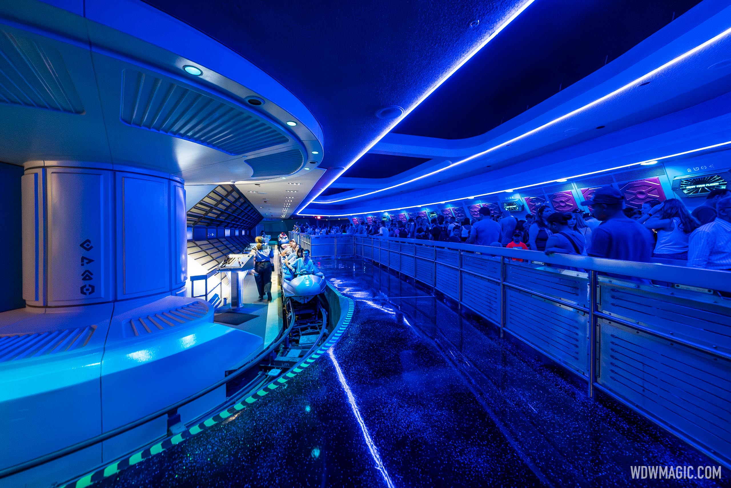 The blue-lit interior boarding station of Space Mountain, with a loaded rocket vehicle at the loading platform, a cast member at a control podium, and a line of guests waiting along the railing under strips of blue light.