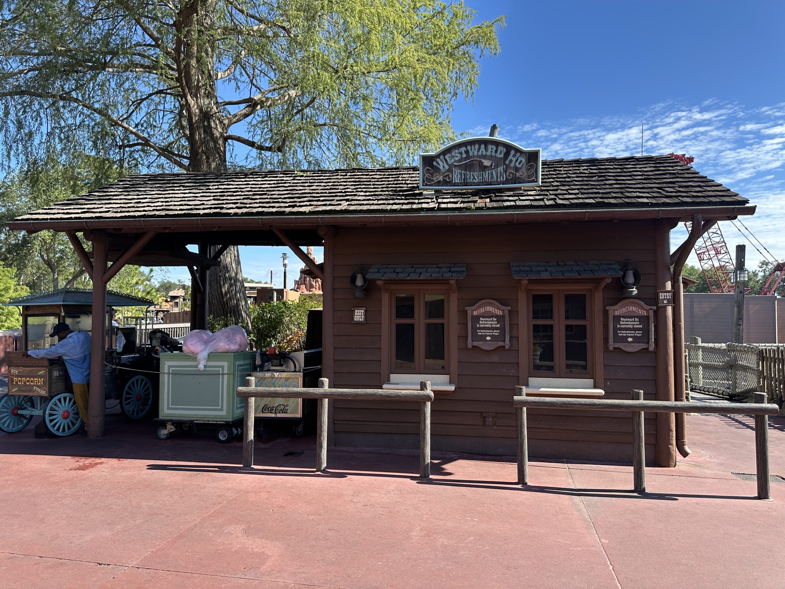 The Westward Ho Refreshments stand intact in Frontierland, a brown wooden building with a shake-shingle roof and its carved sign mounted on the ridge, closure notices on the themed Refreshments boards beside each of the two shuttered windows, a hot popcorn wagon and a cast member at left, and the red lattice boom of a construction crane rising past the right end of the building.
