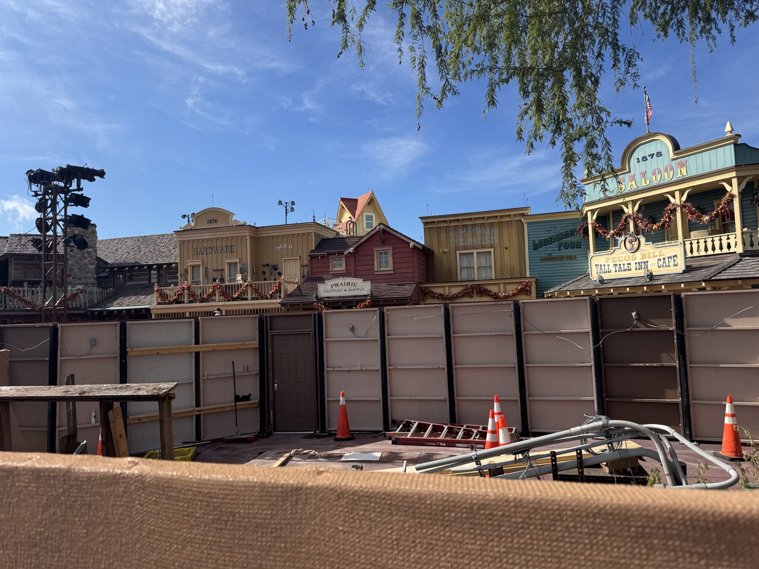 The cleared Westward Ho site photographed over a tan fabric barrier, showing open mauve paving with orange traffic cones, a red extension ladder lying flat and a bundle of gray conduit, with construction wall panels and the Frontierland building facades behind them.