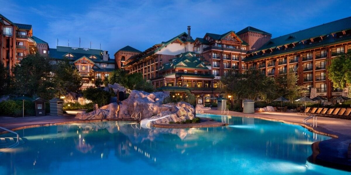 The main Wilderness Lodge building at twilight seen across the large turquoise pool, with green gabled roofs, timber balconies and a rock outcropping at the water's edge.