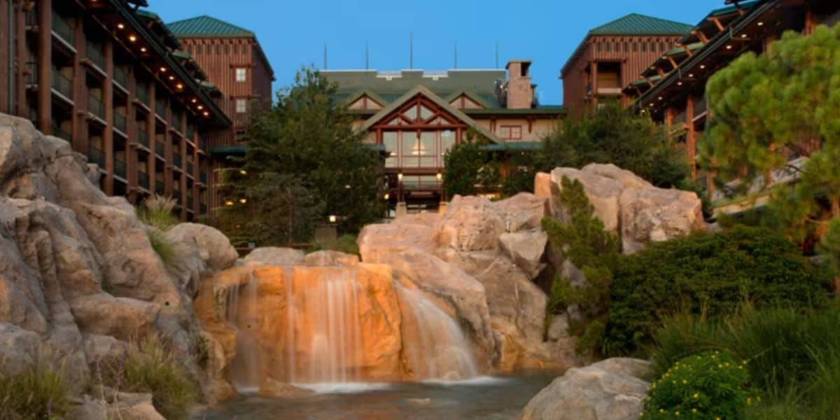 A rock waterfall cascading into a pool in the foreground at dusk, framed by boulders and pines, with the balconied timber wings and gabled main lodge lit behind it.