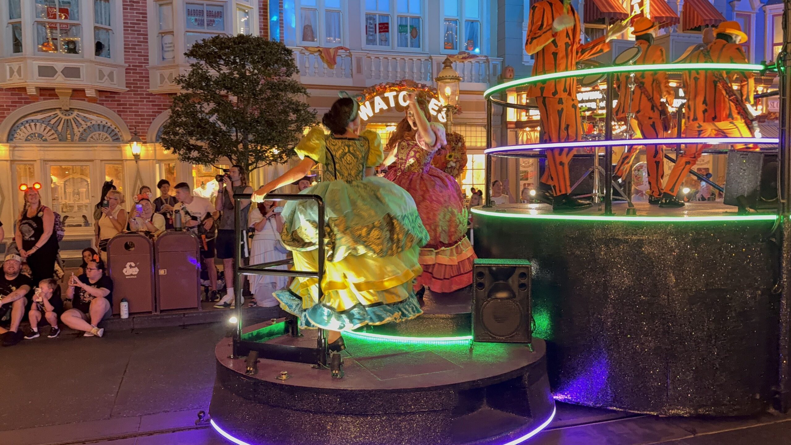 Two parade performers in ruffled ball gowns, one chartreuse and yellow with a teal underskirt, one magenta and coral, dance on a small round platform with a black handrail at the rear of a black glitter float edged in green light. Behind them, performers in orange and black skeleton-print suits play instruments on the float's upper deck, with lit Main Street storefronts and guests filming in the background.