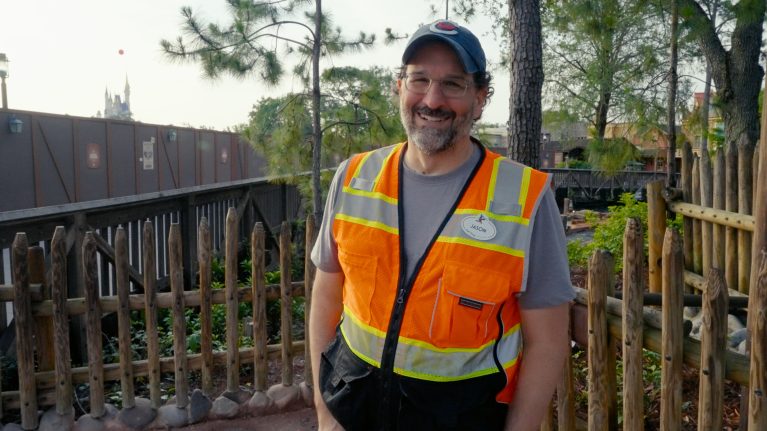 Imagineer Jason Grandt in an orange safety vest on a walkway beside the Piston Peak National Park construction wall, with a small red balloon in the sky behind him and Cinderella Castle in the distance