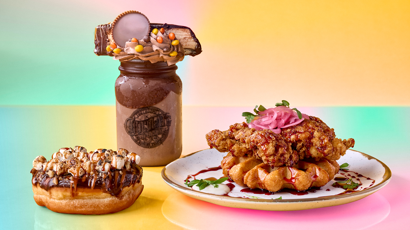 A studio food photo on a pastel rainbow backdrop. At left, a round doughnut topped with chocolate, toasted mini marshmallows, chopped nuts and a caramel drizzle. Center, a mason jar milkshake embossed with the Toothsome Chocolate Emporium logo, topped with chocolate whipped cream, a peanut butter cup, a candy bar and orange and yellow candy pieces. At right, a speckled plate holding a waffle topped with fried chicken, pickled red onion, herbs and a dark red sauce.