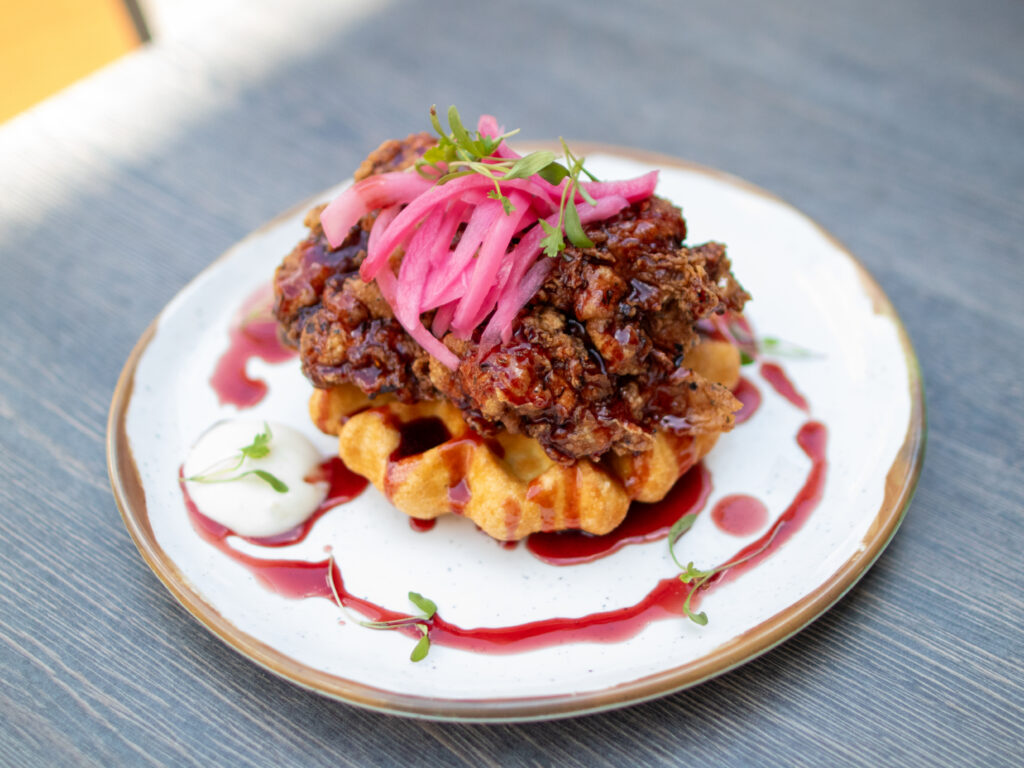 Jerk fried chicken piled on a small round waffle, topped with bright pink pickled onions and micro greens, on a white plate with a red glaze drizzle