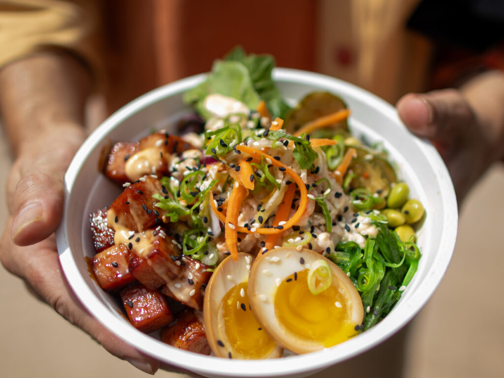 A white bowl held in two hands, filled with coconut rice, glazed cubed spam, a halved soy egg, shredded slaw, edamame, and seaweed salad