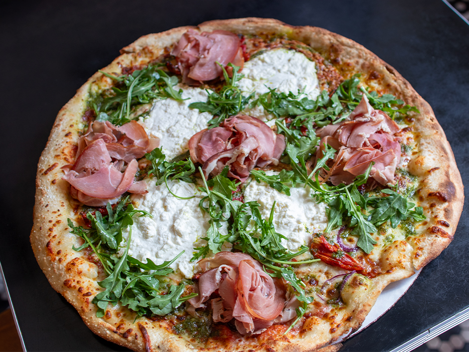 A whole thin crust pizza topped with folded prosciutto, dollops of ricotta, fresh arugula, and pesto