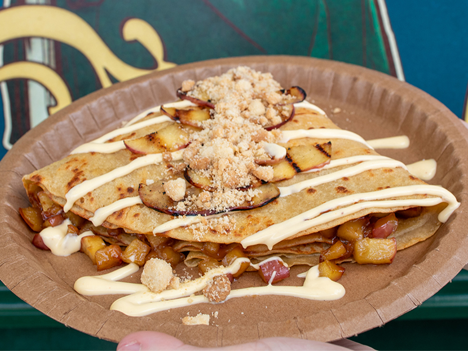 An apple pie crepe on a brown paper plate, folded and topped with grilled red apple slices, brown sugar crumble, and a cream drizzle