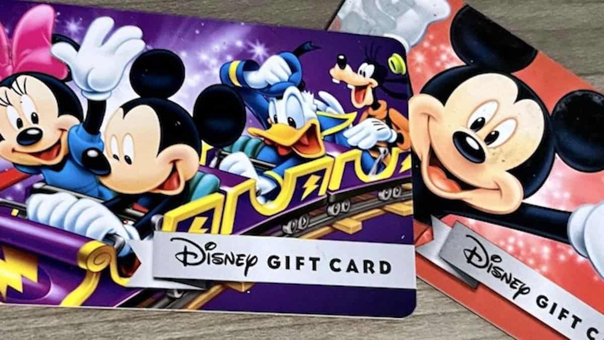Close-up of two physical Disney Gift Cards lying on a wooden surface. The card in front is purple and shows Minnie, Mickey, Donald and Goofy riding a yellow roller coaster train, with a white Disney Gift Card panel across the bottom. Behind it sits a red card with a large Mickey Mouse face and its own Disney Gift Card panel.