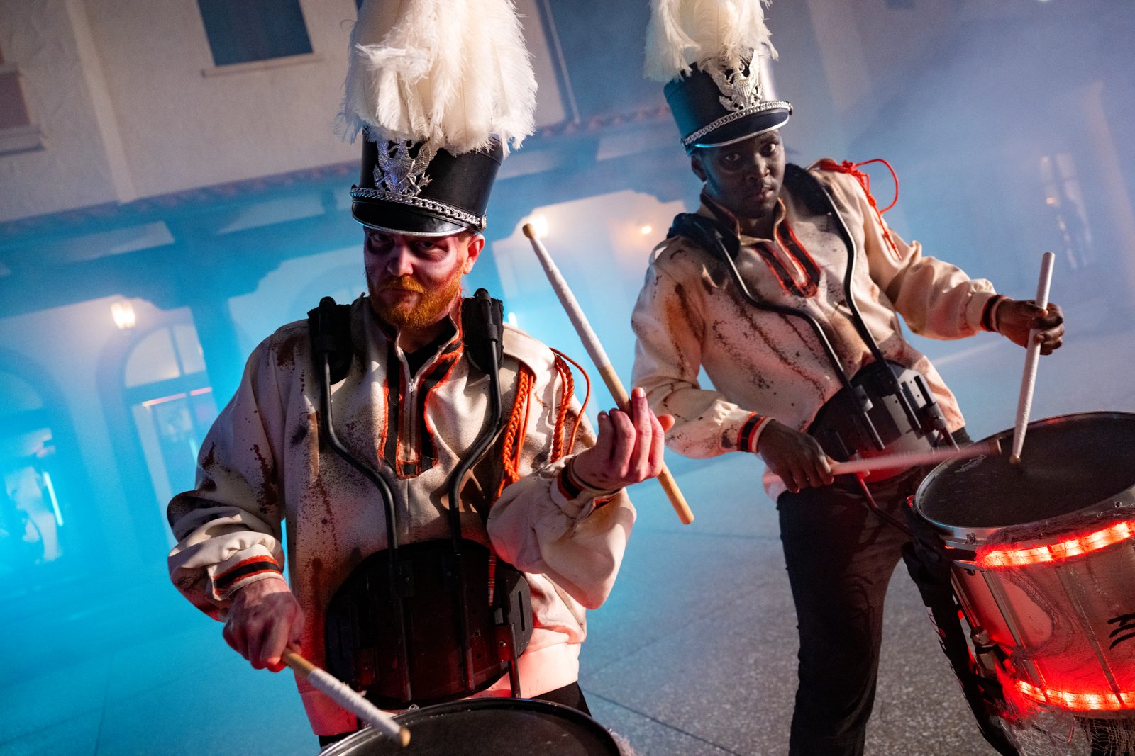 Two drummers in blood-spattered marching band uniforms and tall plumed shakos with corpse makeup play illuminated drums at night