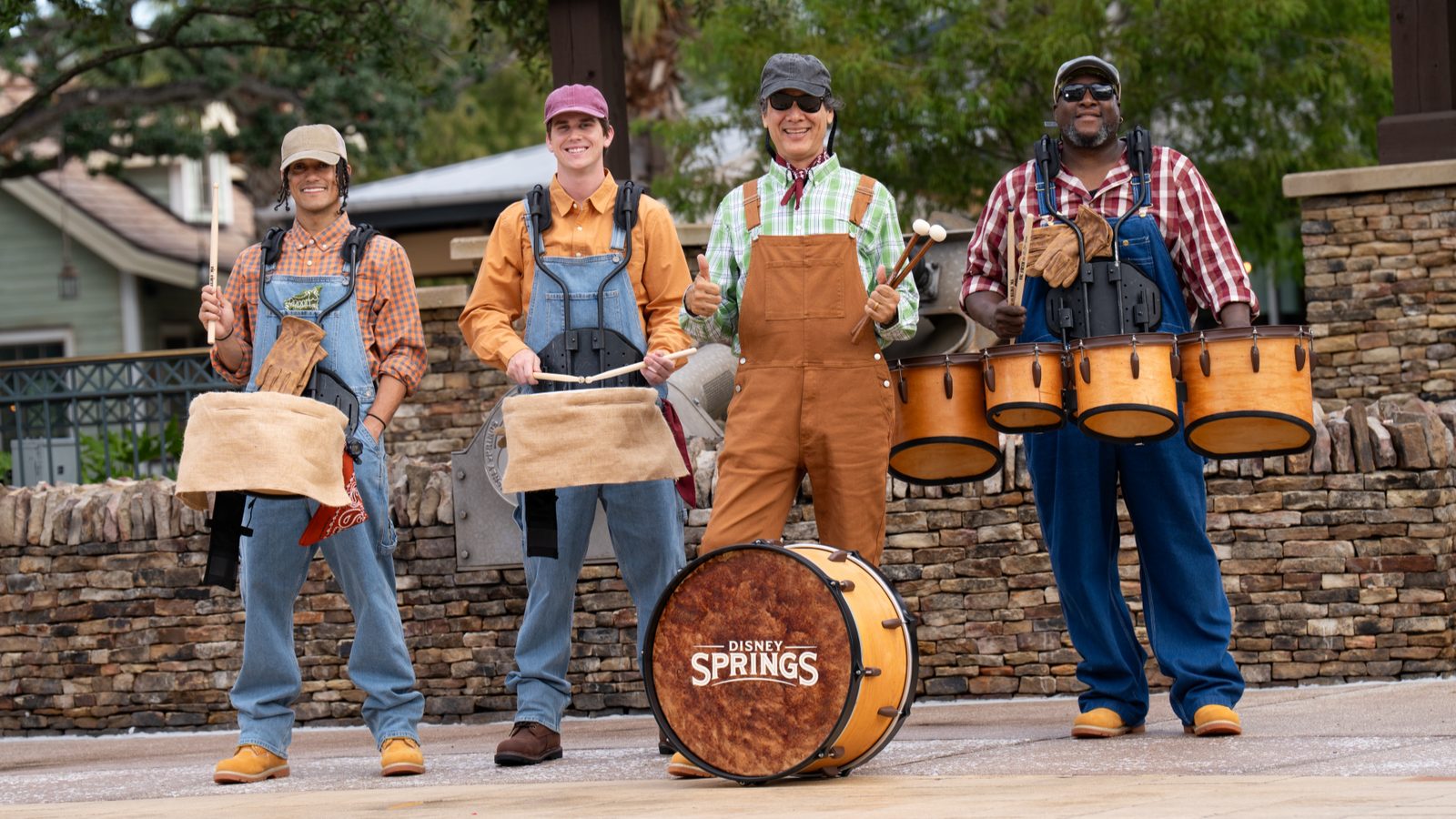 Four Disney Springs performers in denim overalls and plaid shirts stand with burlap-wrapped drums and a bass drum reading Disney Springs