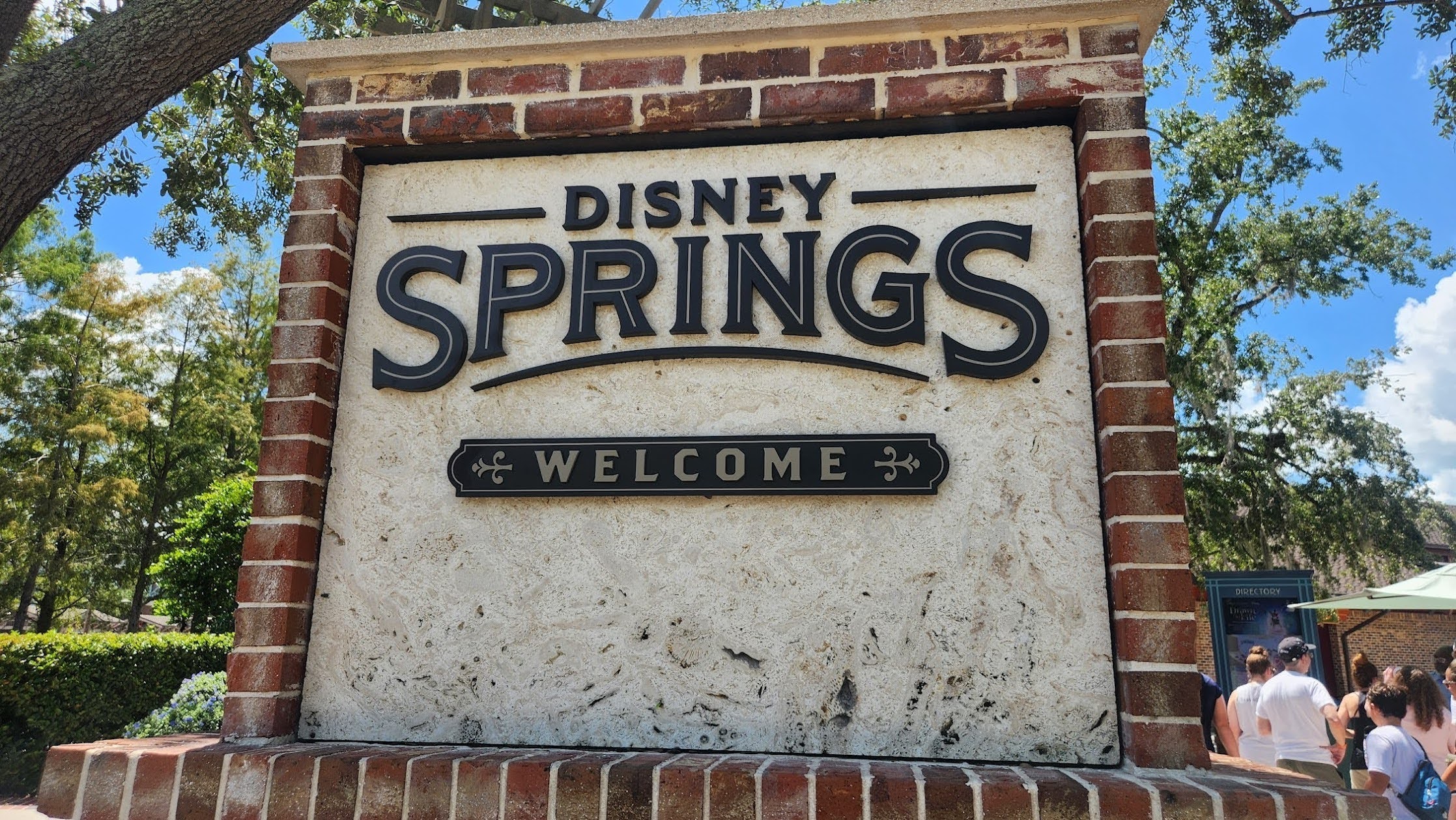 The Disney Springs entrance monument sign on a sunny day, a large stone panel framed in red brick reading DISNEY SPRINGS in dark serif lettering above a smaller WELCOME plaque. Oak branches hang over the top of the frame, and a directory kiosk with guests walking past it is visible at the right edge.