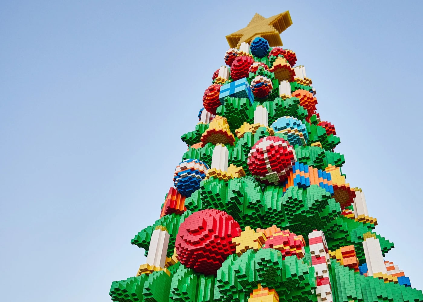 Looking steeply up at a Christmas tree built entirely from LEGO bricks against a clear pale blue sky. The green brick branches are hung with oversized brick ornaments in red, blue and white, plus wrapped presents and candy canes, and a chunky yellow brick star sits on top.