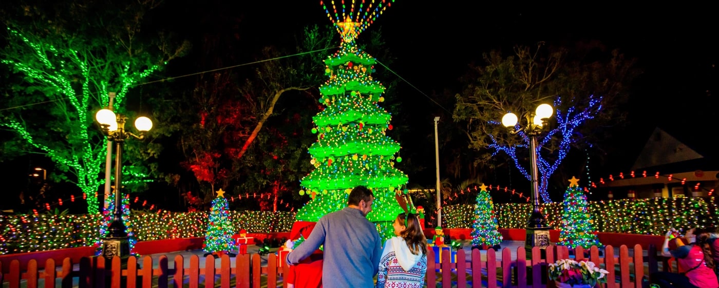 A nighttime park scene. A tall tiered Christmas tree glowing green under thousands of lights stands at center, hung with round red, yellow and green ornaments beneath a lit yellow star topper with strings of colored bulbs fanning upward and outward from it, with smaller lit trees around it and hedges packed with multicolored lights. Trees at left are wrapped in green lights and trees at right in blue. A man and a young girl in a reindeer headband stand with their backs to the camera behind a low red picket fence.