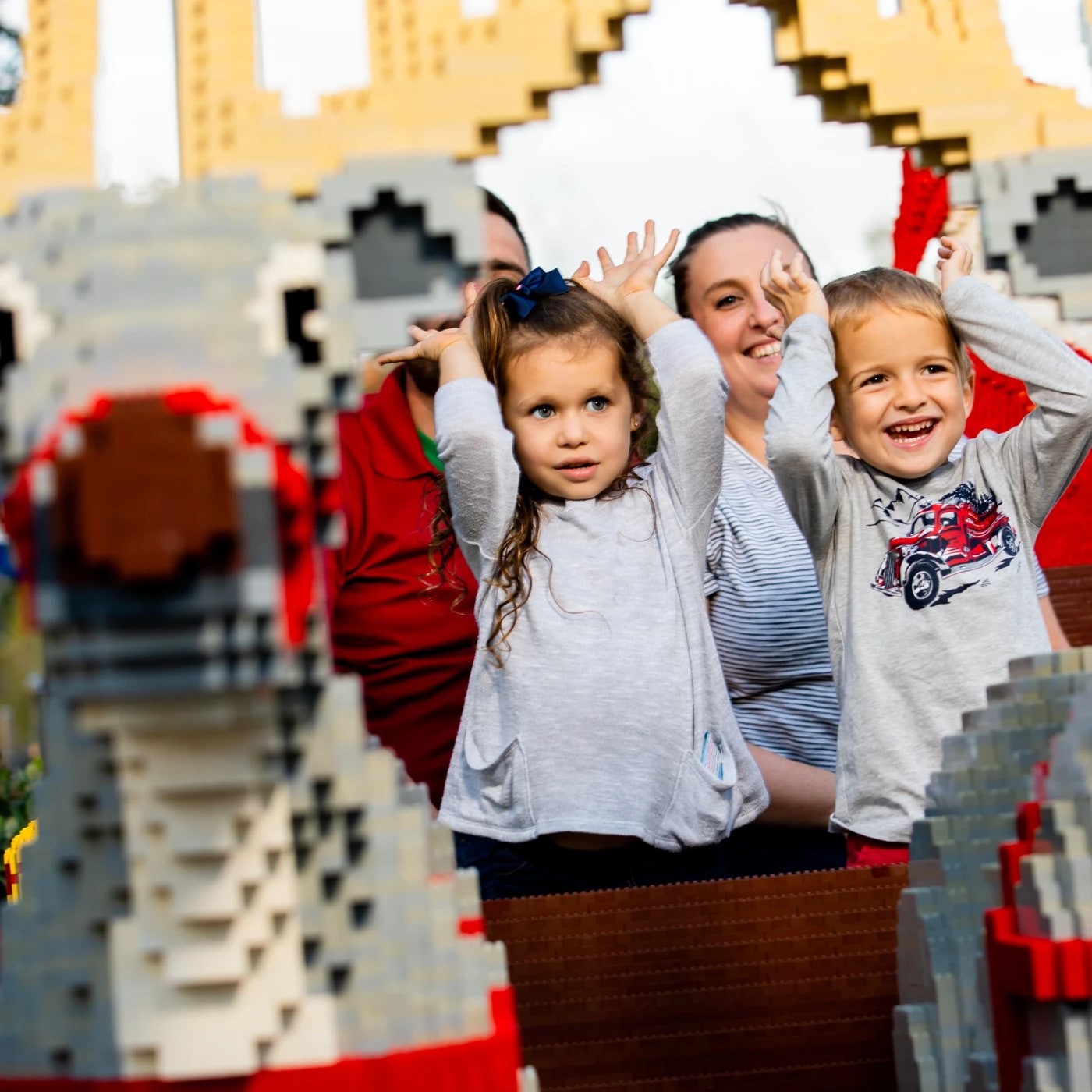 Two young children stand in a sleigh built from LEGO bricks with their hands raised beside their heads like antlers, the boy grinning wide and the girl looking straight at the camera. Two adults stand behind them, the nearer one a smiling woman in a striped shirt making the same antler gesture. In the blurred foreground is the head of a large brick built reindeer with a dark red nose, and tan brick antlers spread across the top of the frame.