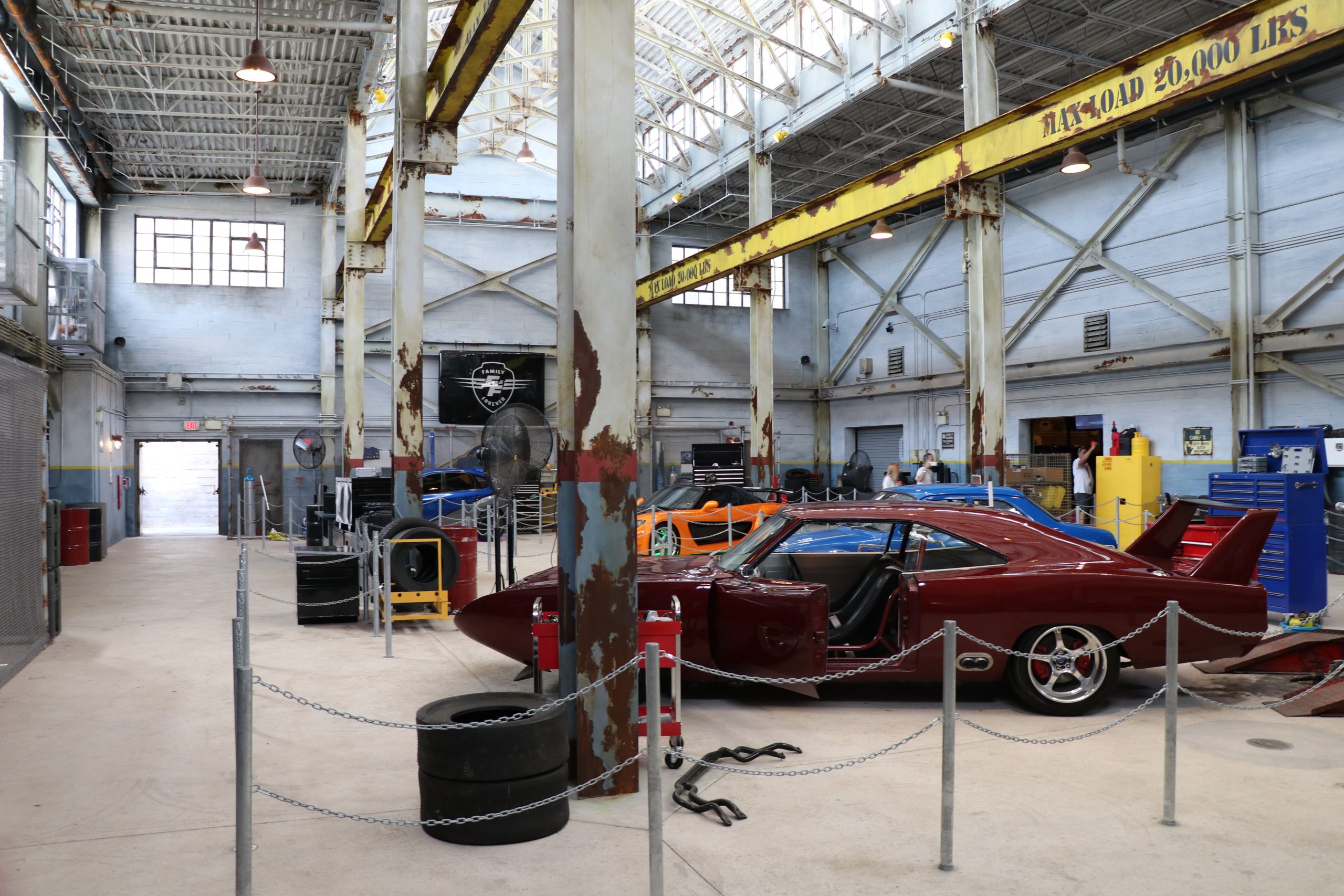 The Fast and Furious Supercharged queue garage with a maroon hero car, its door open, stacked tires in the foreground and a few guests at the back of the room