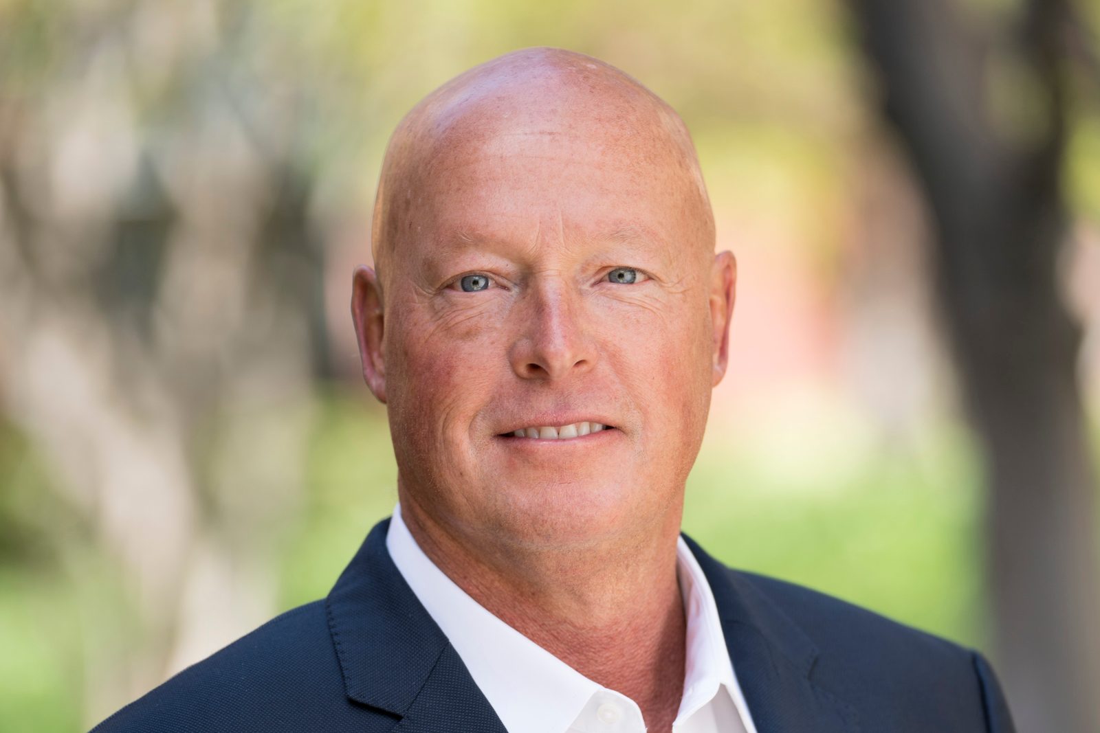 A corporate portrait of Bob Chapek, bald and clean shaven, in a navy blazer and open collared white shirt against a blurred green background