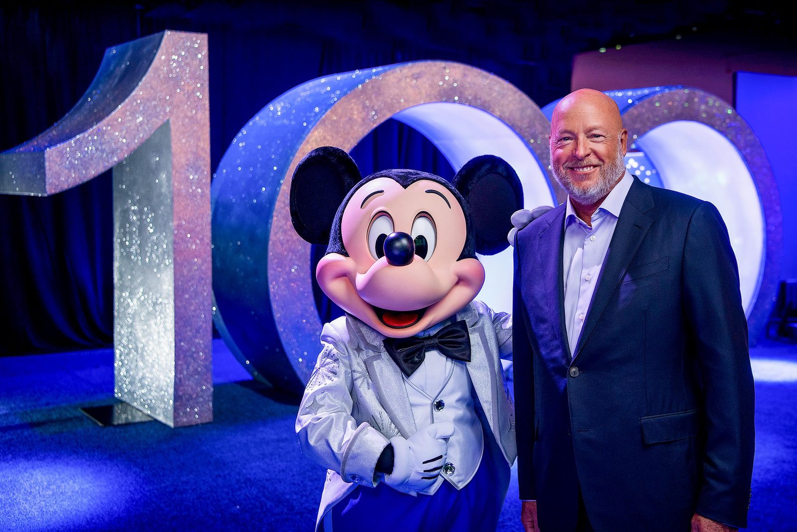 Bob Chapek in a dark suit standing beside Mickey Mouse in a silver tuxedo in front of large glittering numerals reading 100 under blue stage light