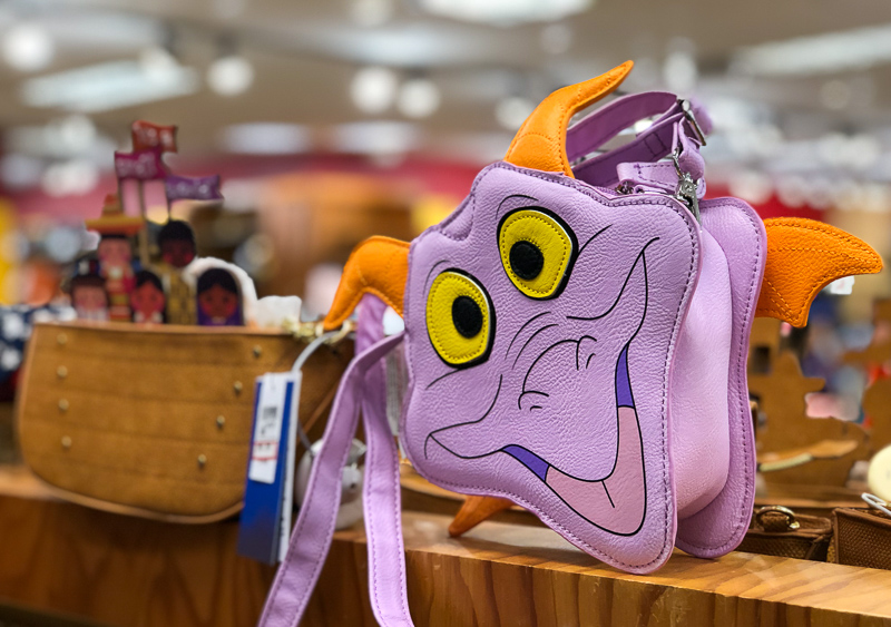 A lavender Figment shaped crossbody bag with yellow eyes and orange horns and wings sits on a wooden shelf inside a store