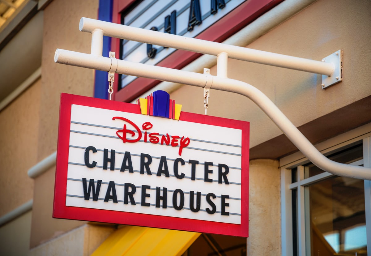 A hanging red and white marquee sign reading Disney Character Warehouse outside an outlet store, topped with a small blue, red and yellow crest
