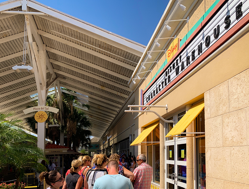 Guests gather along a sunlit outlet mall walkway outside a Disney Character Warehouse storefront with yellow awnings