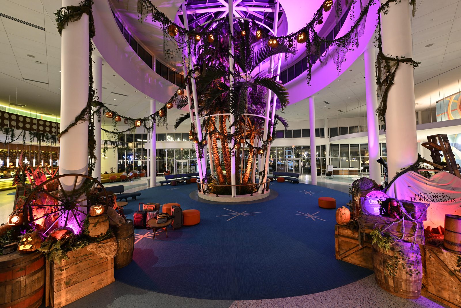 A hotel lobby atrium under purple light with a tall palm at its center, strings of glowing jack-o-lanterns and mossy garland hung between white columns, and wooden barrels and crates around the edges