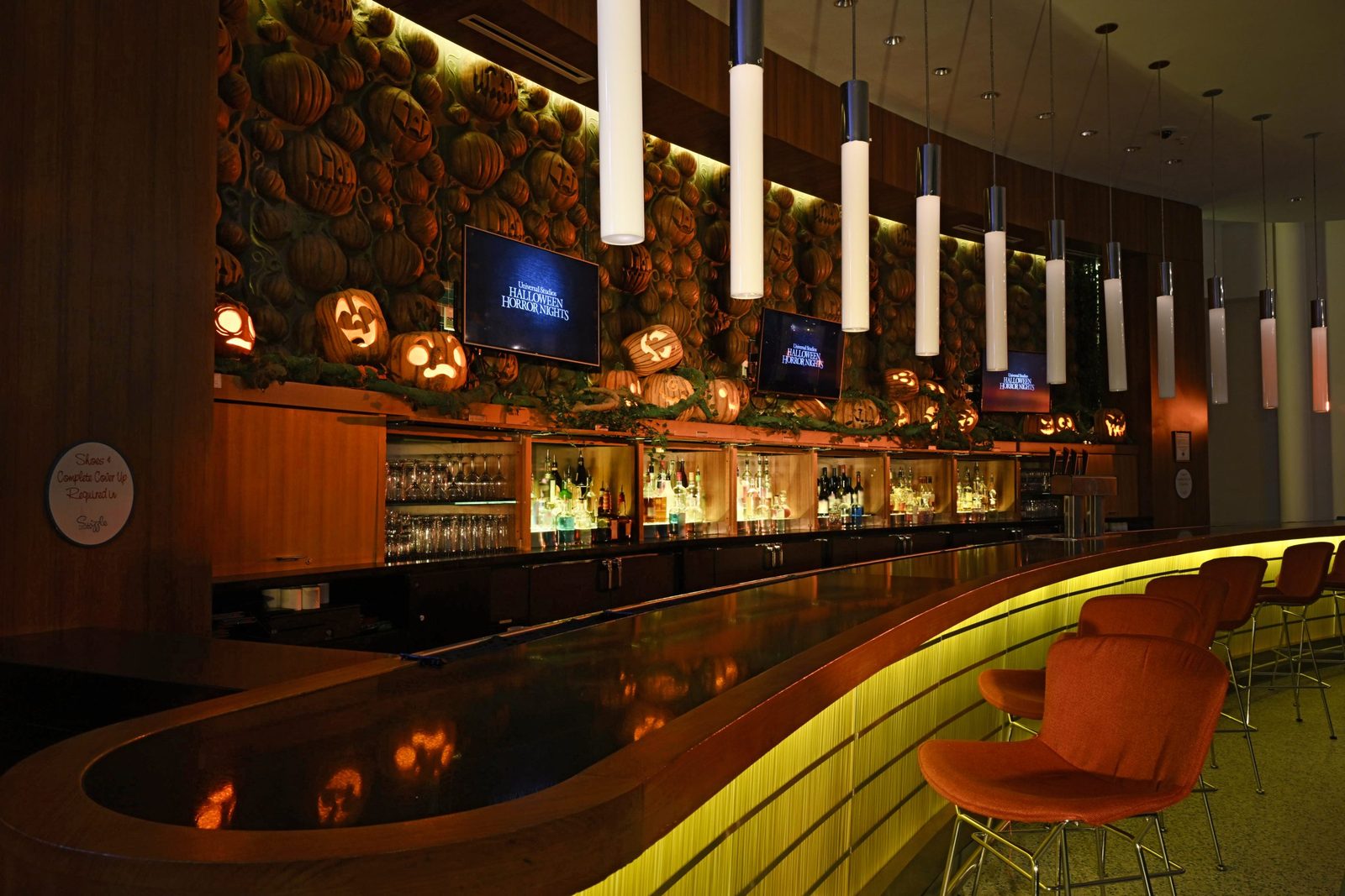 A long curved wooden bar under a wall packed with carved and glowing jack-o-lanterns, with three screens reading Halloween Horror Nights and orange chairs at right