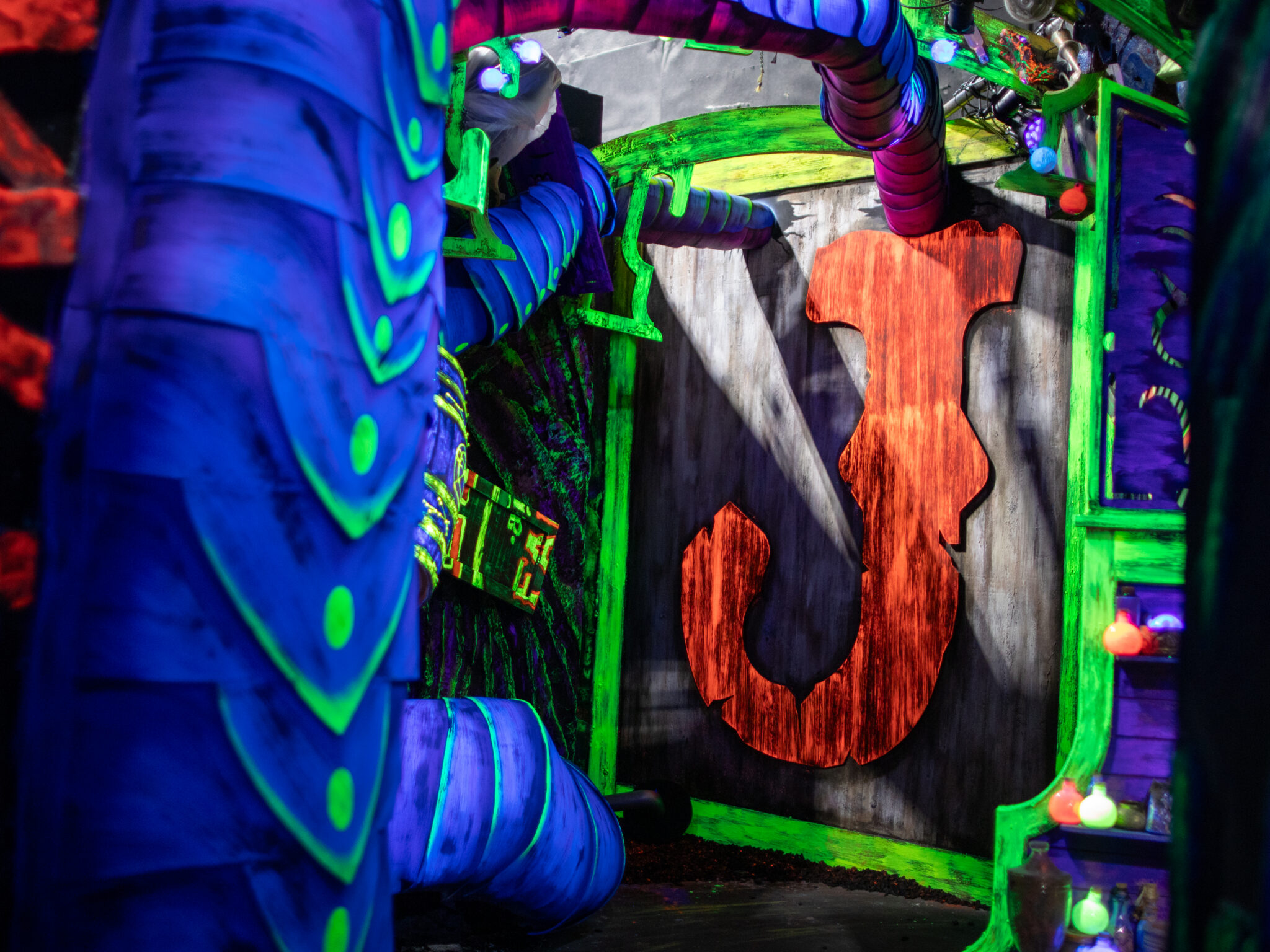 A blacklit room where a large red letter J is mounted on a wooden wall framed by glowing green trim, segmented tentacles and shelves of luminous bottles