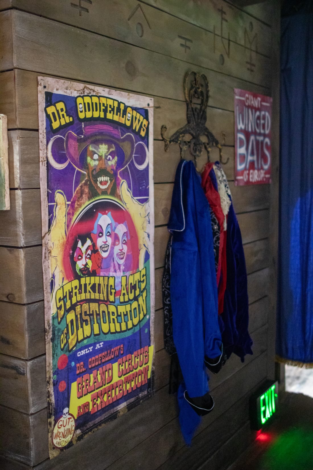 A weathered wooden wall inside the house holding a vintage style poster reading DR. ODDFELLOWS STRIKING ACTS OF DISTORTION, only at Dr. Oddfellow's Grand Circus and Exhibition. Costumes hang on an iron hook beside it and a smaller sign reads GIANT WINGED BATS OF EUROPE.