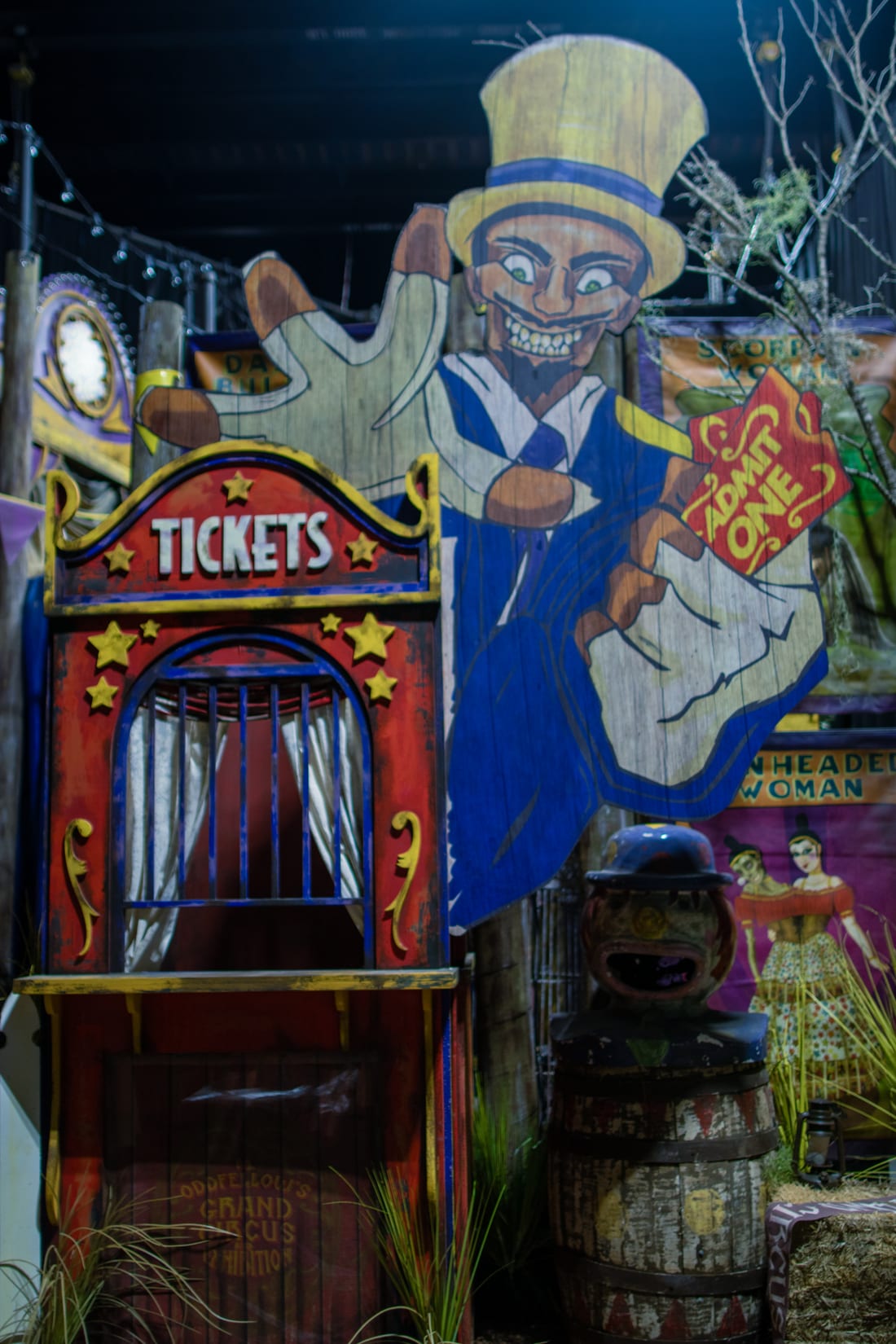 The house entrance, styled as a carnival midway. A wooden cutout of Dr. Oddfellow in a yellow top hat and blue suit leans out over a red ticket booth marked TICKETS, holding an ADMIT ONE ticket, with sideshow banners behind him.