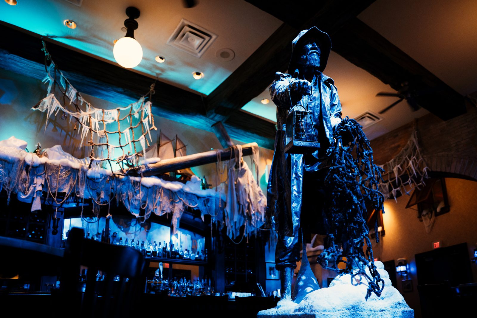 A lantern carrying figure in a long coat and wide brimmed hat standing on a mound of snow inside a bar, holding a tangle of frozen rope, with iced netting and a wooden ship's spar lit blue behind