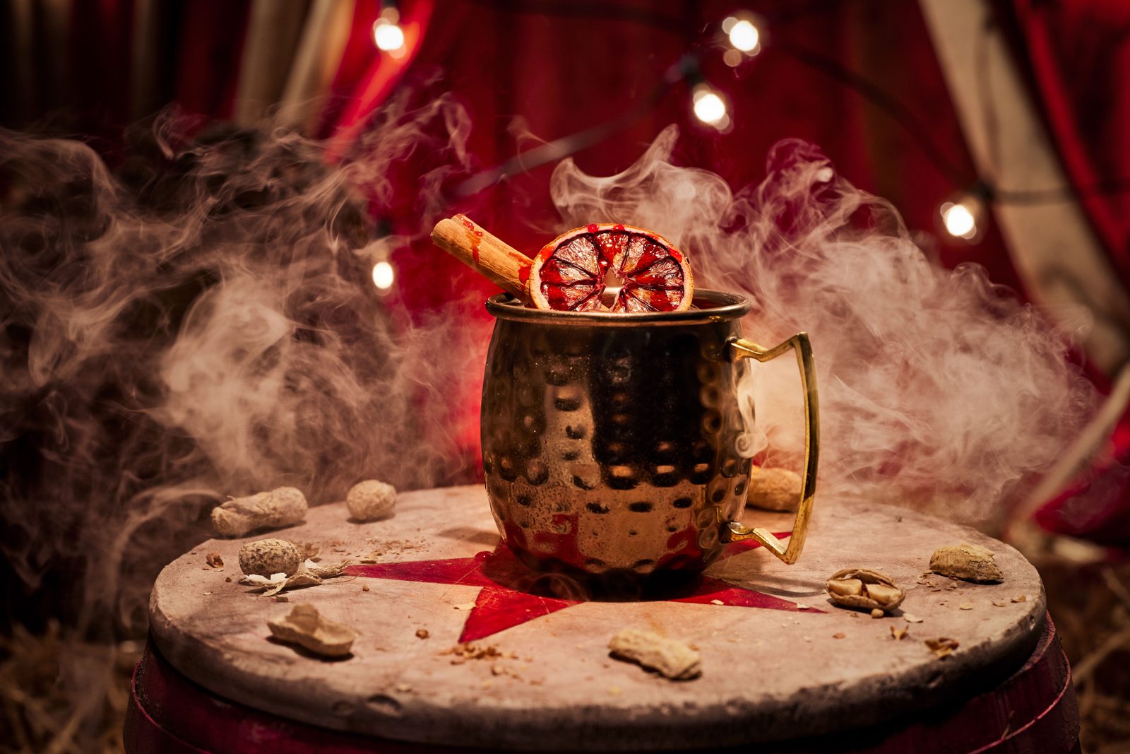 A copper mug wreathed in white smoke holding a drink garnished with a dried citrus wheel and a cinnamon stick, sitting on a barrel top scattered with peanut shells against a red striped tent backdrop