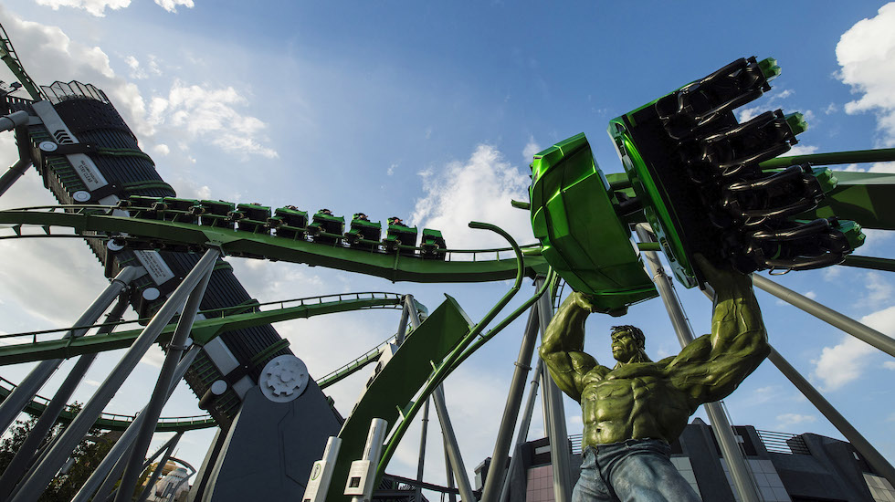A train of riders on the green track of The Incredible Hulk Coaster beside a statue of the Hulk holding a coaster car above his head, with the launch tower at left under a blue sky