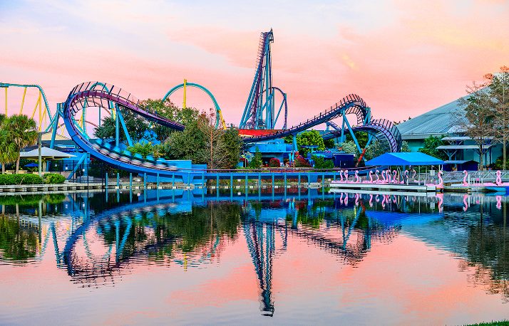 A wide view across a lagoon at sunset with several coaster layouts silhouetted against a pink sky and mirrored in still water, with flamingos on the far bank