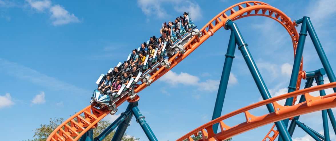 A coaster train packed with riders on bright orange track supported by teal columns, curving against a blue sky with scattered cloud