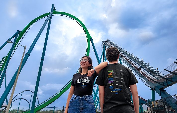 Two people in front of a green coaster and a lift hill, one facing the camera in a shirt reading National Roller Coaster Day 2026 and one turned away showing a printed list of coaster names and stats on the back