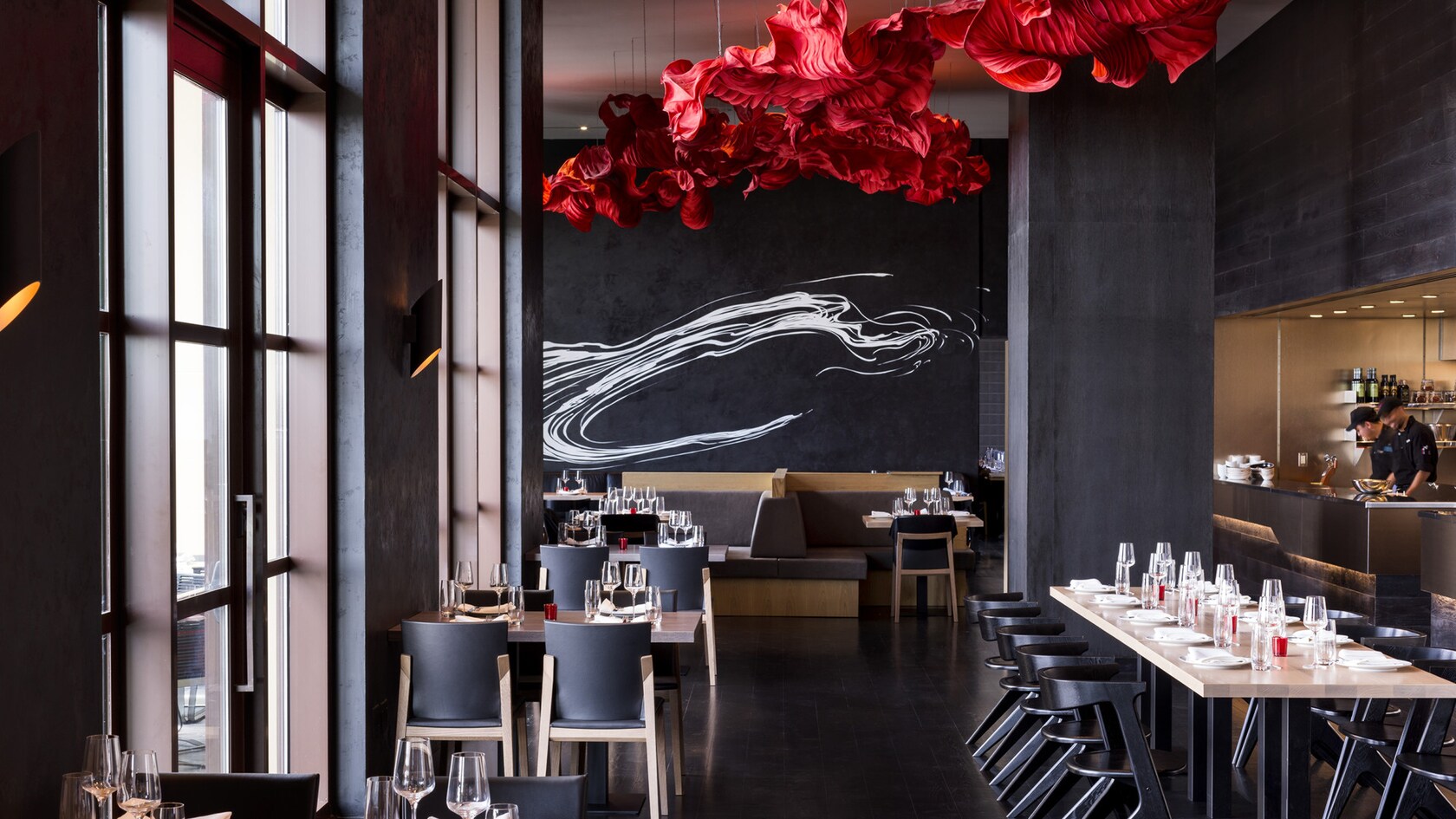 The dining room at Capa, with floor to ceiling windows along the left wall, dark grey walls, a large sculptural red fabric installation hanging from the ceiling, a white brushstroke mural on the back wall, and an open kitchen at right where two chefs are working.