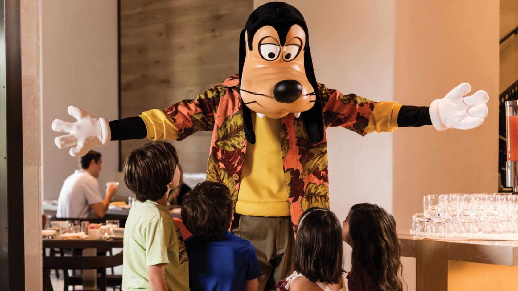 Goofy in a floral shirt over a yellow shirt, arms spread wide, greeting four children in a bright restaurant dining room, with another guest seated at a table behind and stacked glassware on a counter at right.