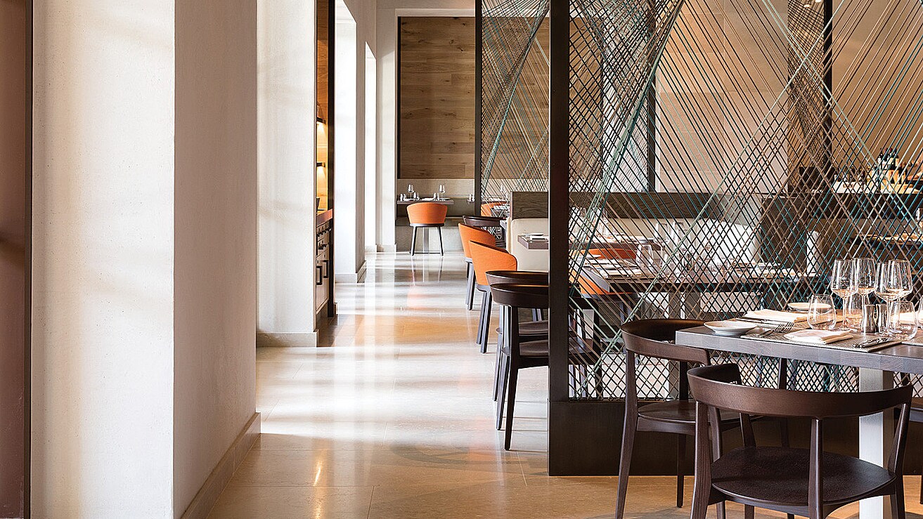 The dining room at Ravello, a bright sunlit space with pale stone floors and columns, dark wood chairs, orange upholstered chairs, and a decorative crosshatched cord screen dividing the room, with tables set with wine glasses.