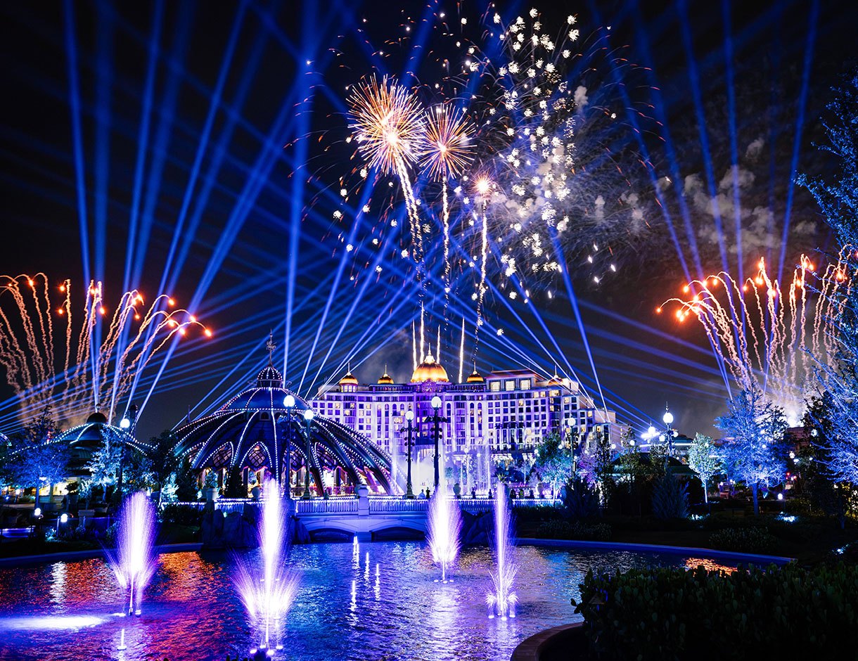 Night view of Celestial Park with fireworks bursting overhead, blue searchlight beams fanning across the sky, lit fountains in a reflecting pool and a domed glass pavilion in front of a large illuminated hotel