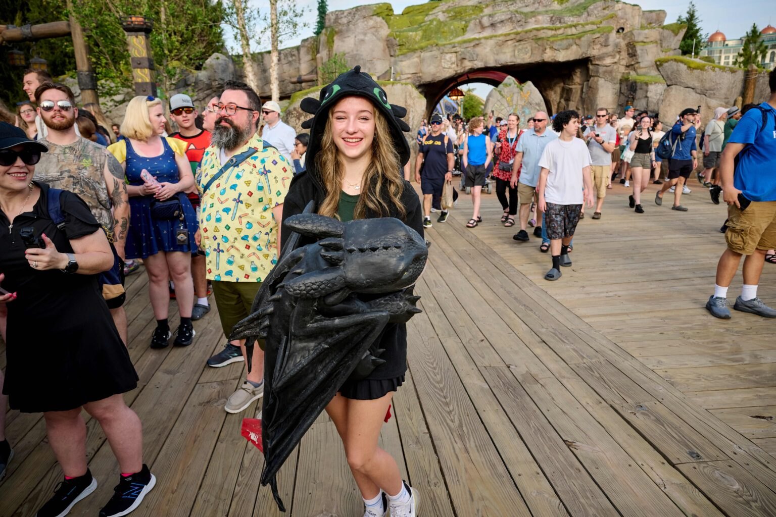 A dense daytime crowd on a wide wooden boardwalk in How to Train Your Dragon - Isle of Berk, with a girl in a black Toothless hooded jacket carrying a large Toothless plush in the foreground and a stone archway behind