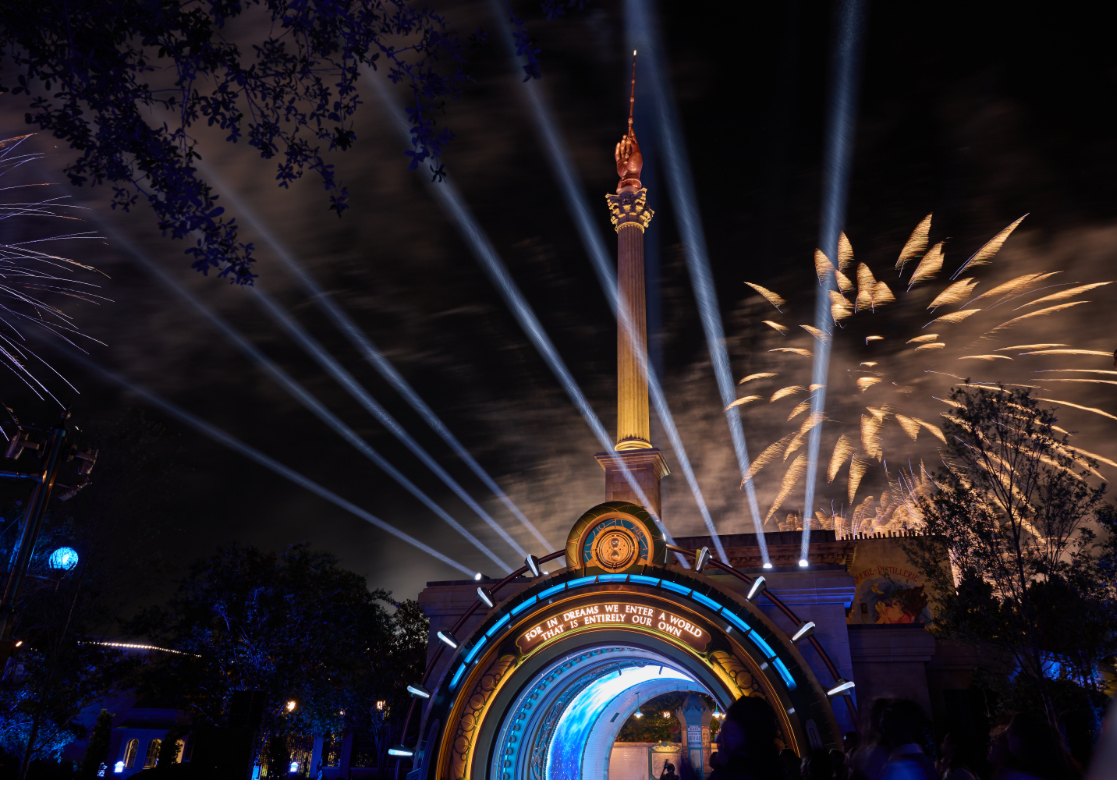 The lit archway into The Wizarding World of Harry Potter - Ministry of Magic at night, ringed in blue light with a tall golden column topped by a hand holding a wand, searchlight beams and gold fireworks overhead