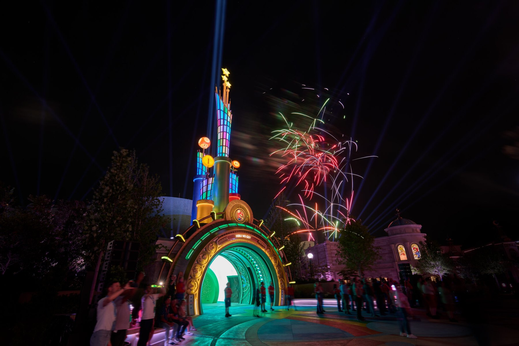 The illuminated warp pipe entrance arch to SUPER NINTENDO WORLD at night, glowing green and gold and topped by gold stars, a Super Mushroom, a coin and a Mario medallion, with fireworks and searchlights behind it and guests gathered in front
