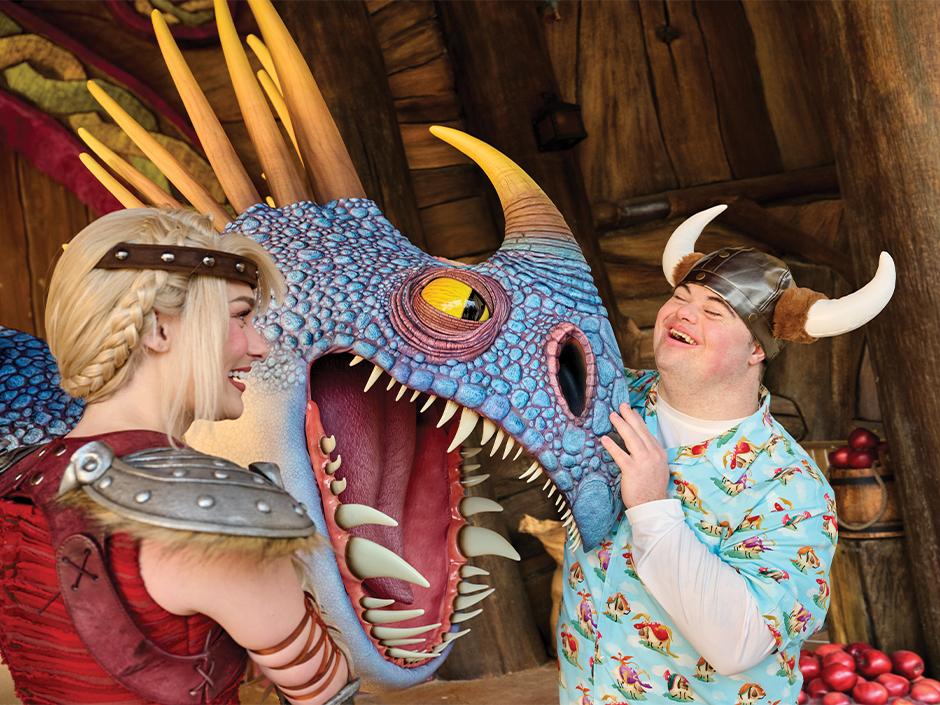 Astrid in Viking costume at left facing a large blue scaled dragon puppet with its mouth open, while a guest in a horned helmet laughs and touches its snout