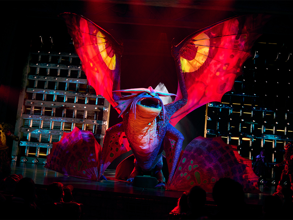 A large blue and purple dragon puppet on a dark stage with two wings raised high and two more fanned low, all four lit deep red, standing above silhouetted audience heads