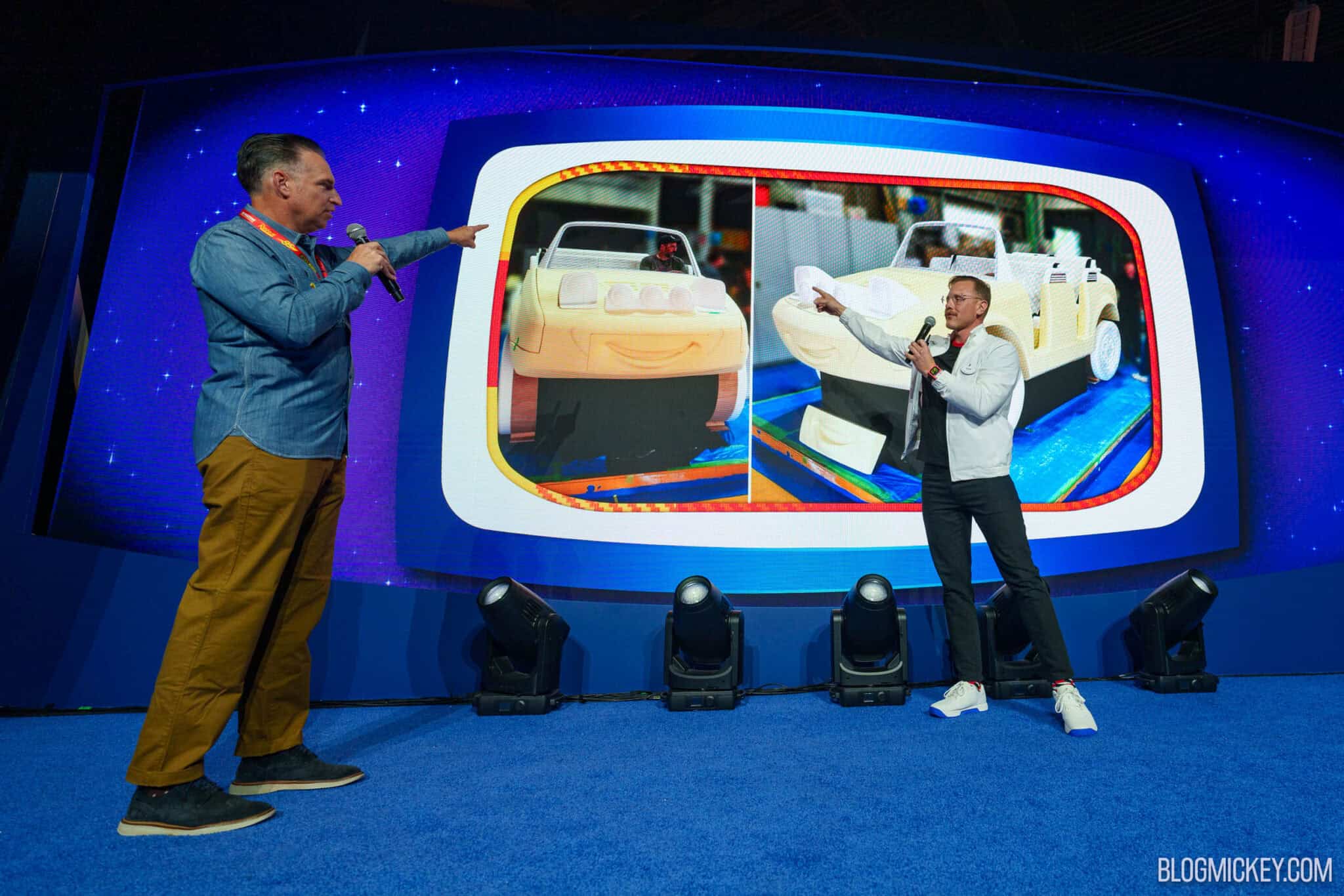 Two presenters on a stage point at a large screen showing two photos of an unpainted cream foam car