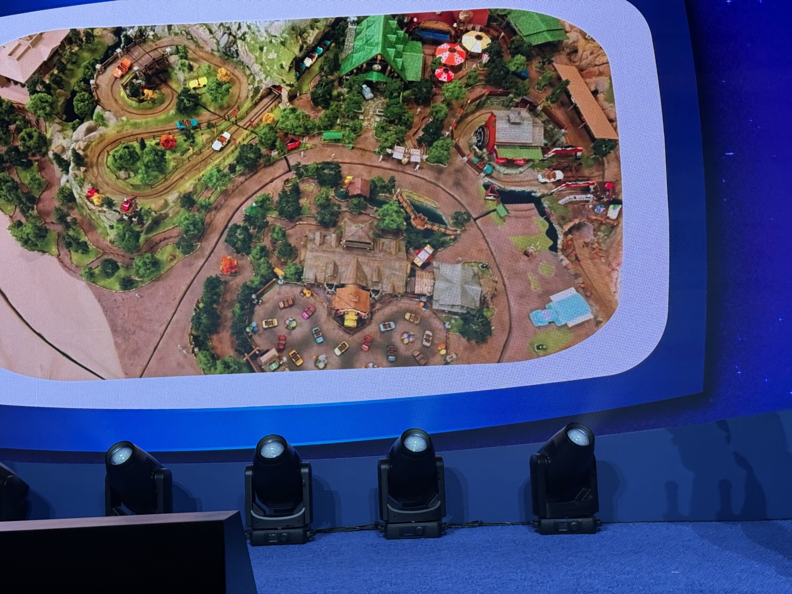 Presentation screen showing an overhead photo of a detailed scale model, with a winding dirt track at upper left and a large dirt arena scattered with tiny cars at lower center
