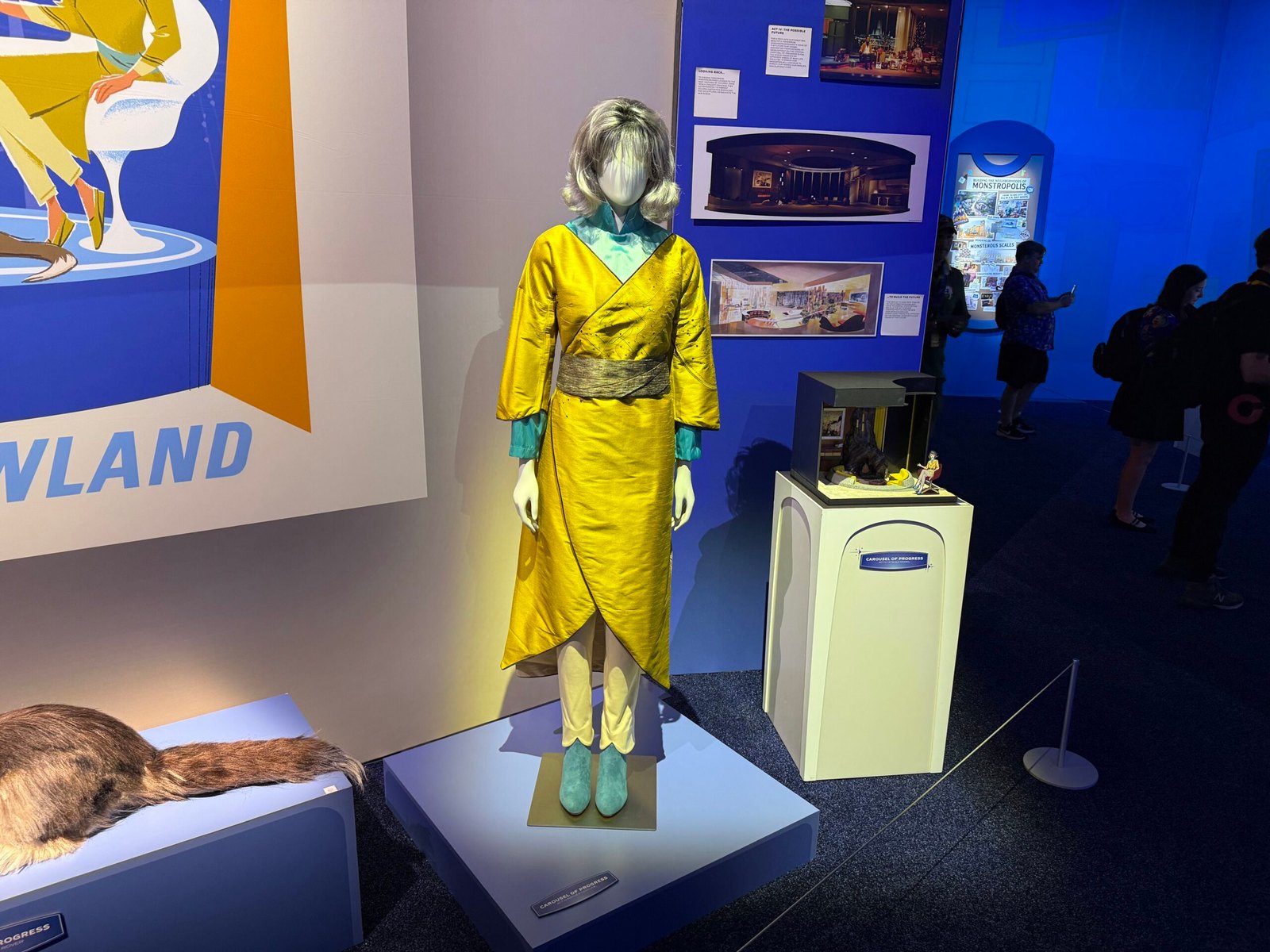 A gold wrap dress with teal collar and cuffs displayed on a mannequin, with a lit model case to the right