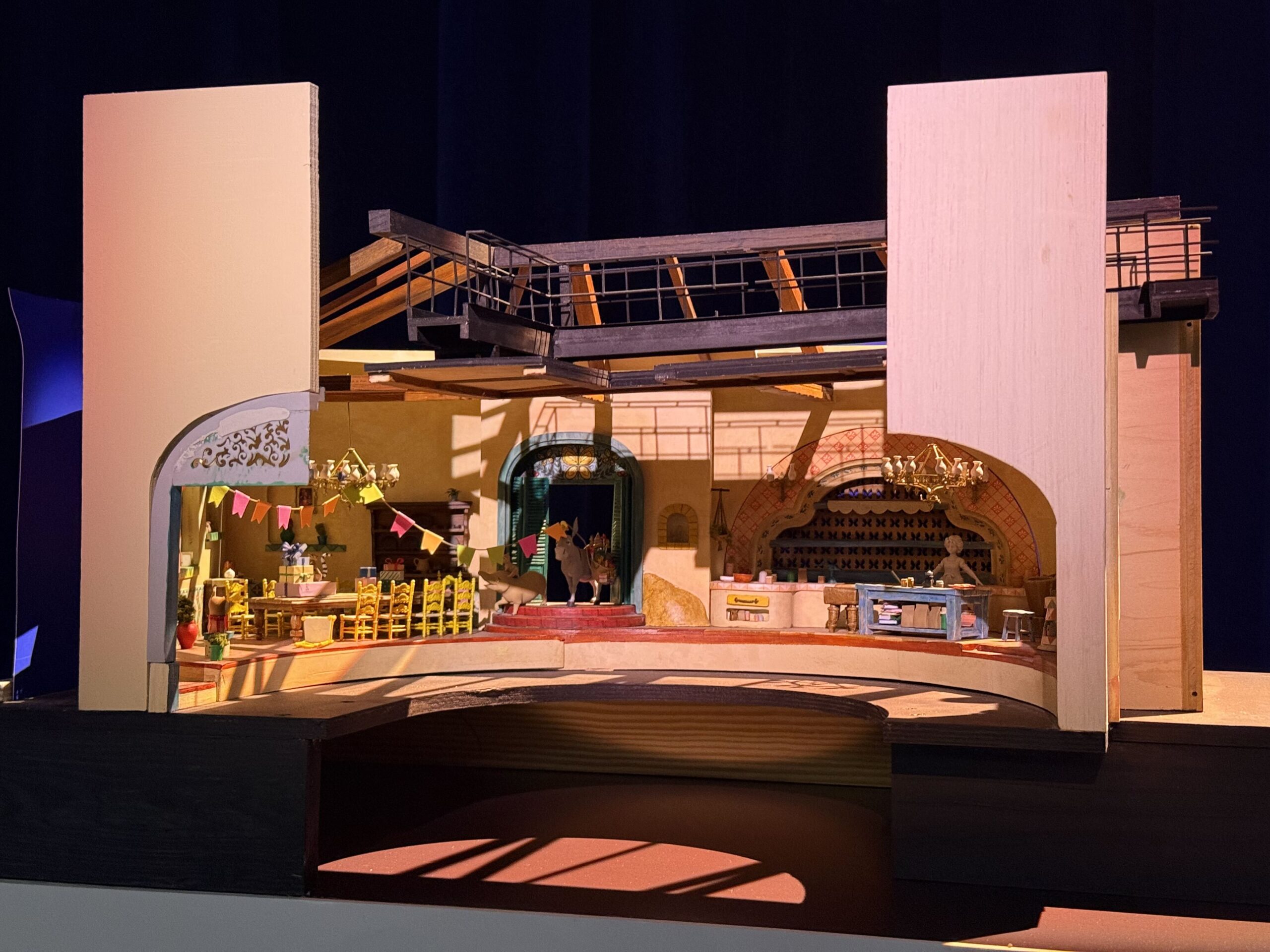 A cutaway scale model of an interior scene showing a dining room with yellow chairs and a table of wrapped presents under colorful bunting on the left, a teal arched doorway with a donkey and a pig in the center, and a kitchen with a blue prep table on the right, with the model's exposed roof trusses and catwalk above.