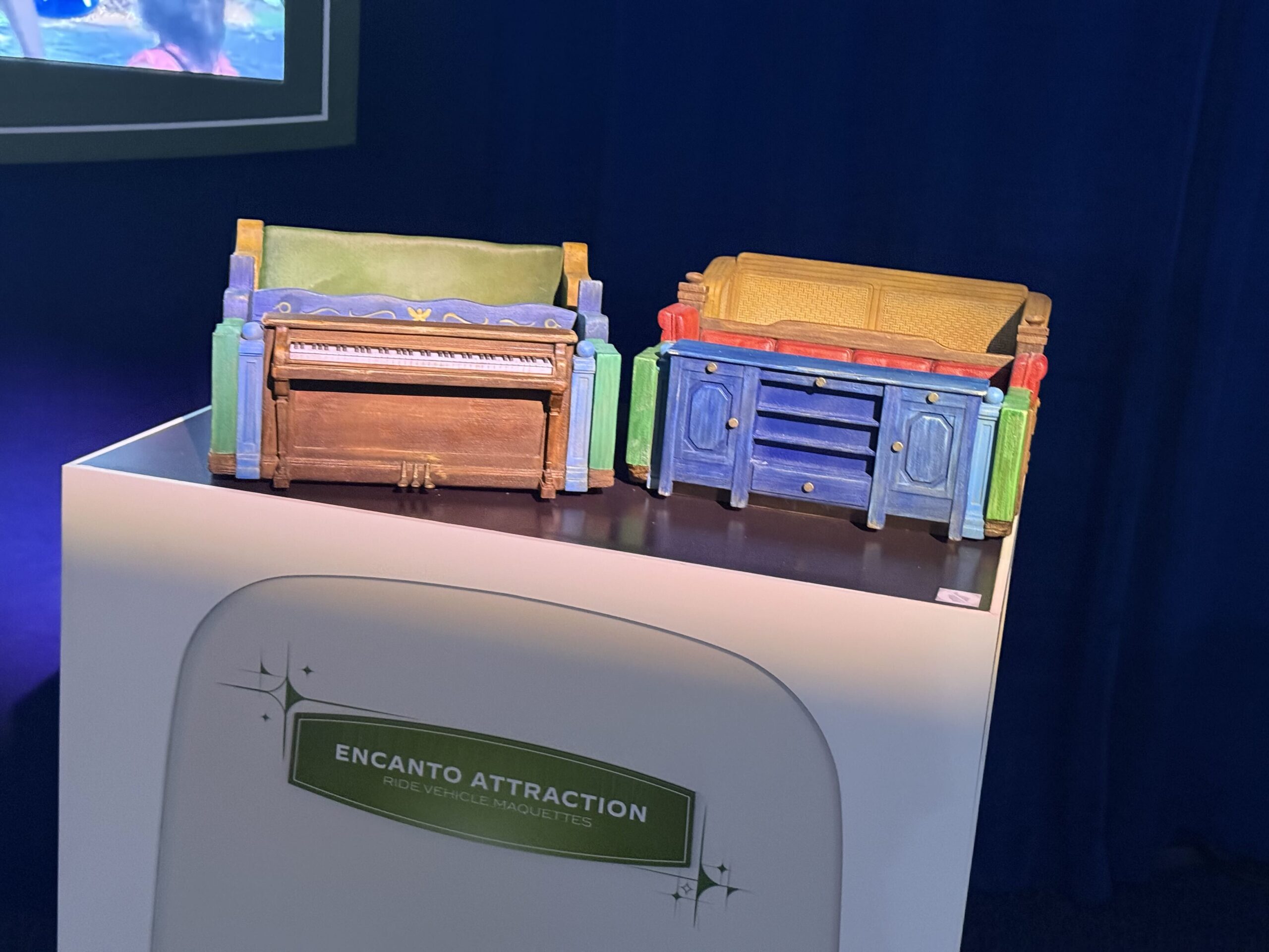 Two colorful ride vehicle models on a white plinth. The left model has an upright piano as its front with a green padded bench behind it, the right has a blue dresser with brass knobs and a woven golden bench, and the plinth is labeled Encanto Attraction Ride Vehicle Maquettes.